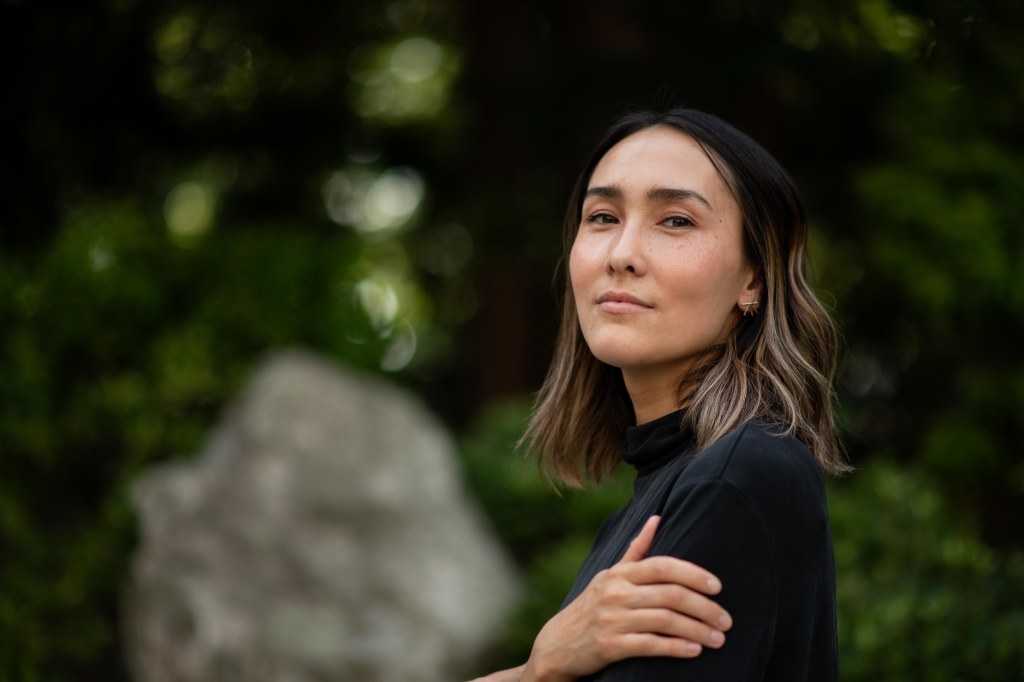 A close-up portrait of a woman with long, wavy hair, wearing a black top, standing outdoors with soft green foliage in the background.