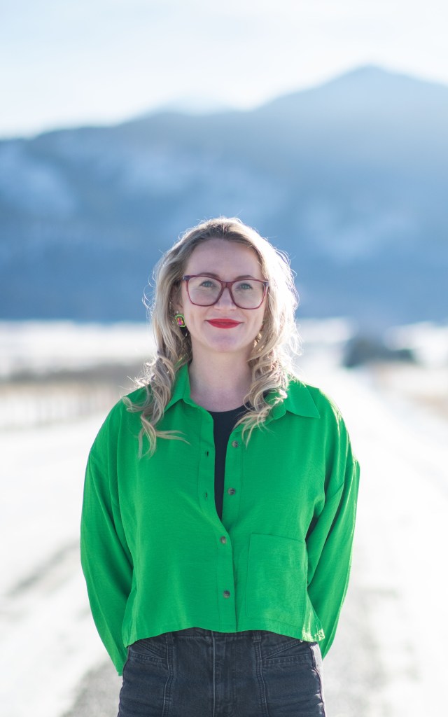 A woman with wavy blonde hair stands on a road surrounded by mountains, wearing a green button-up shirt and glasses.