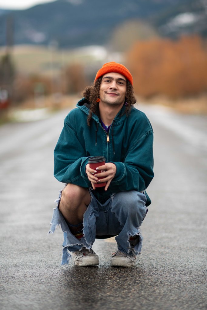 A young man squatting on a road, holding a coffee cup, wearing a teal hoodie, orange beanie, and distressed denim jeans.