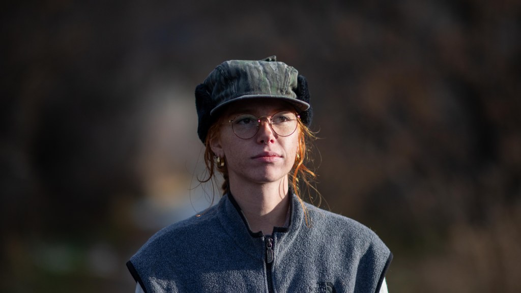 Close-up of a woman with red hair wearing a gray vest and a forest green cap, standing outdoors with a blurred background.