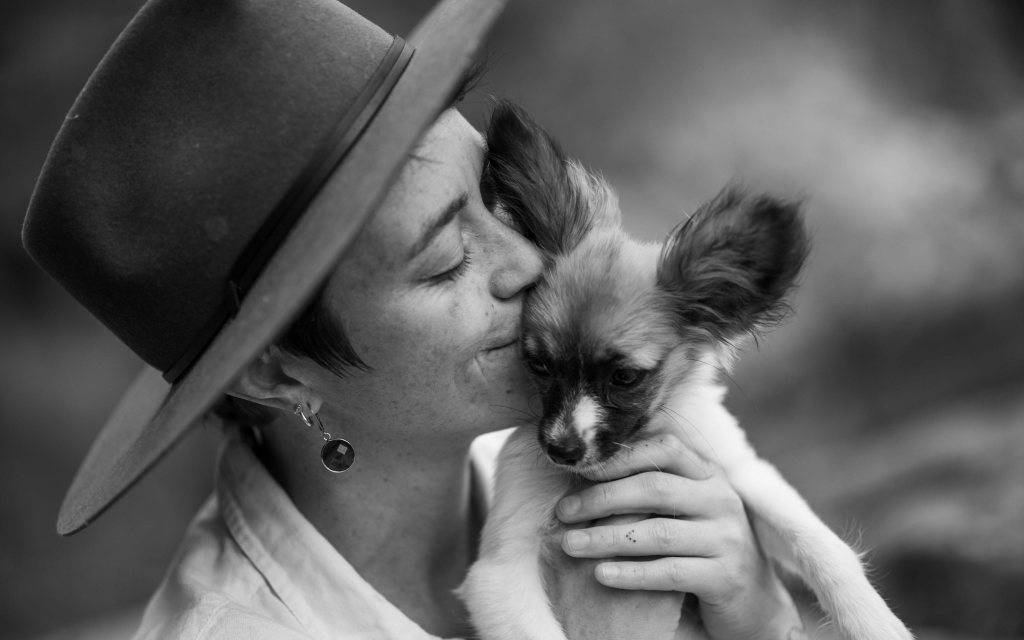 A woman wearing a hat lovingly kisses her small, fluffy dog in a black and white setting.