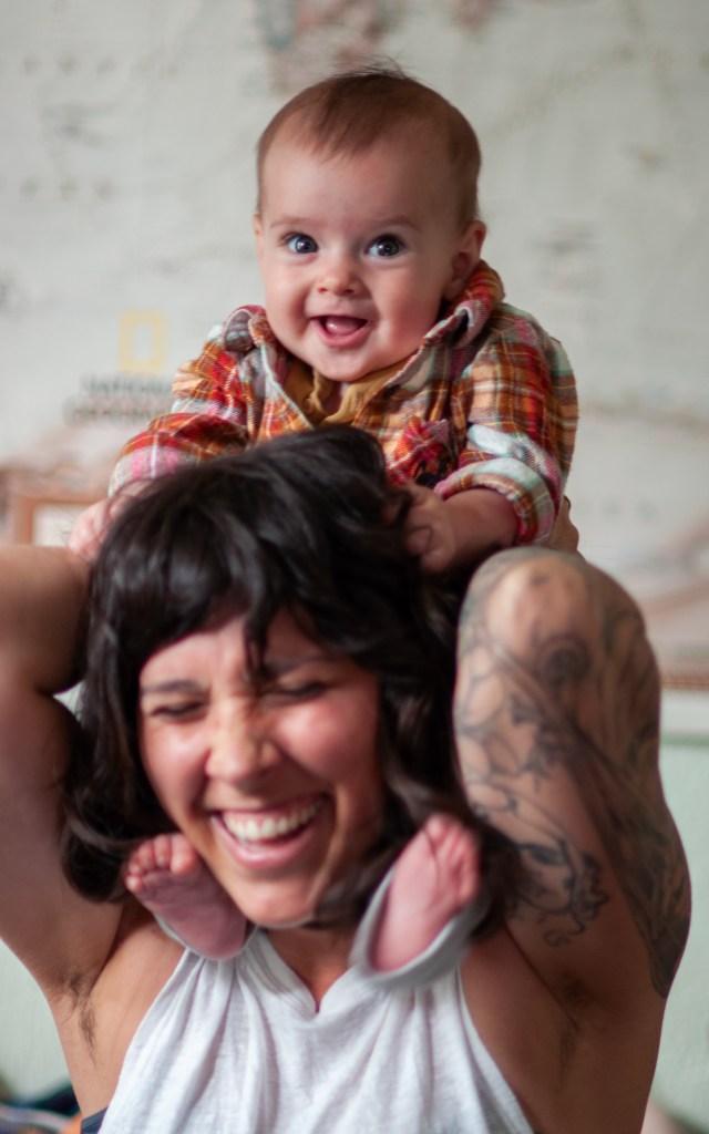 A joyful woman laughing while holding a baby on her shoulders, both with happy expressions, against a blurred background.